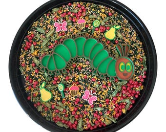 May include: A round black tray filled with a sensory mix of red, yellow, and green dried grains and pasta shapes. Centred is a large green caterpillar figure with a red face. Surrounding it are small colourful icons of butterflies, cupcakes, lollipops, and pears.