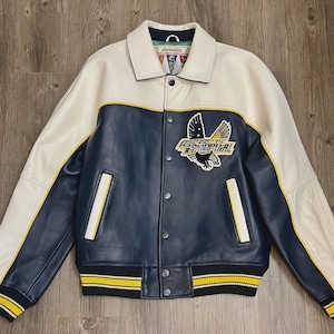 May include: A leather varsity jacket in cream, navy blue, and yellow. The jacket features a cream-colored collar and sleeves, a navy blue body, and yellow stripes on the cuffs and hem. An embroidered eagle patch is on the chest.