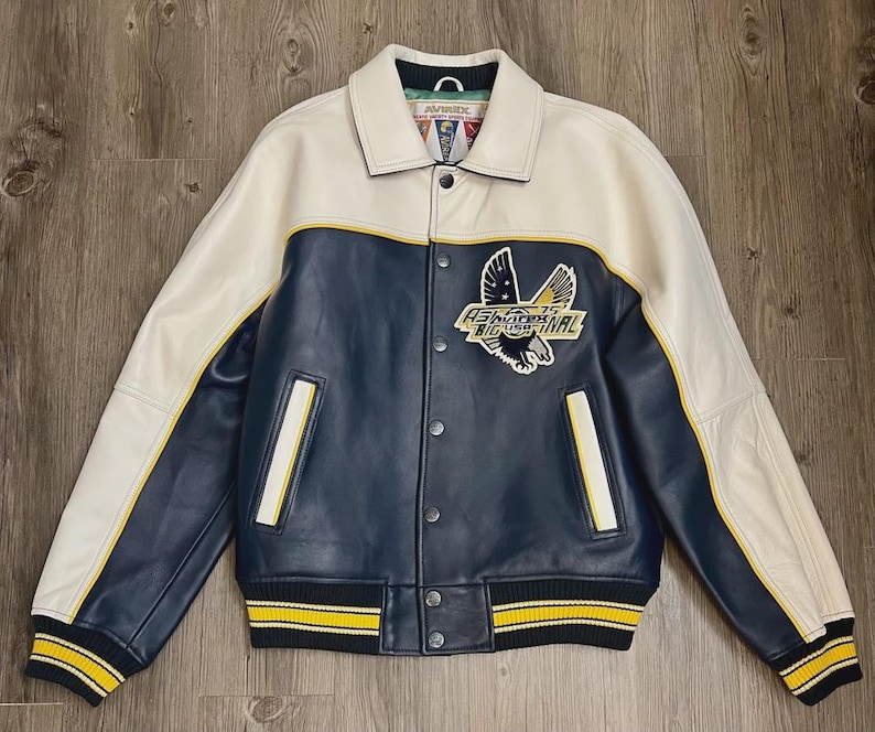 May include: A leather varsity jacket in cream, navy blue, and yellow. The jacket features a cream-colored collar and sleeves, a navy blue body, and yellow stripes on the cuffs and hem. An embroidered eagle patch is on the chest.