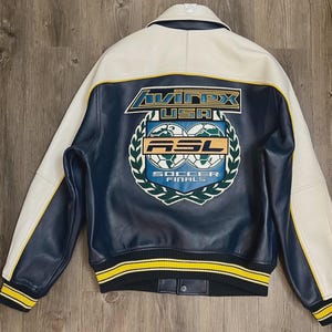 May include: A navy blue and white leather jacket with yellow accents. The back features a large emblem with the text "Avirex USA" and "ASL Soccer Finals". The jacket has a ribbed collar, cuffs, and hem.