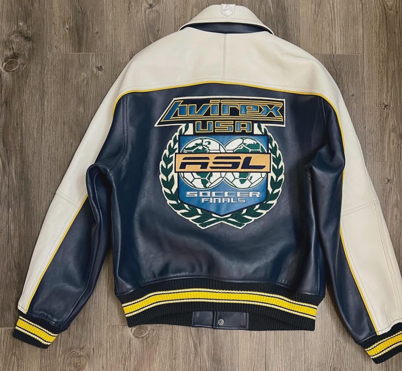 May include: A navy blue and white leather jacket with yellow accents. The back features a large emblem with the text "Avirex USA" and "ASL Soccer Finals". The jacket has a ribbed collar, cuffs, and hem.