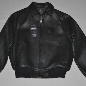 May include: Black leather bomber jacket with a zip-up front, ribbed cuffs, and a collar. The jacket features a small emblem on the chest and a tag with text. A classic outerwear piece.