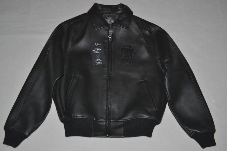 May include: Black leather bomber jacket with a zip-up front, ribbed cuffs, and a collar. The jacket features a small emblem on the chest and a tag with text. A classic outerwear piece.