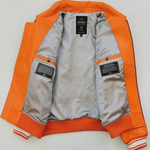 May include: Open orange leather jacket with a gray satin lining. The jacket features a ribbed orange and white striped hem and cuffs. The interior has two pockets and a label that reads "AVIREX LEATHER".