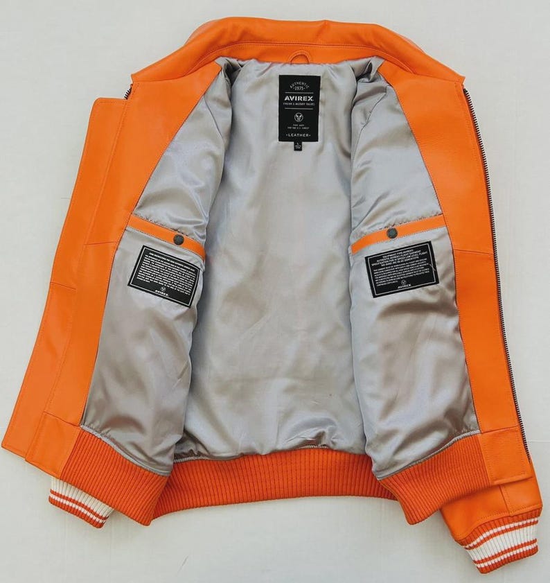 May include: Open orange leather jacket with a gray satin lining. The jacket features a ribbed orange and white striped hem and cuffs. The interior has two pockets and a label that reads "AVIREX LEATHER".