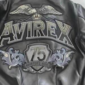 May include: Black leather jacket with embroidered patches. The back features a large eagle with outstretched wings above the brand name "AVIREX". Below the name is a shield with the number "75" and the text "AVIATOR'S KING".
