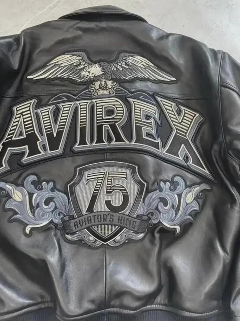 May include: Black leather jacket with embroidered patches. The back features a large eagle with outstretched wings above the brand name "AVIREX". Below the name is a shield with the number "75" and the text "AVIATOR'S KING".