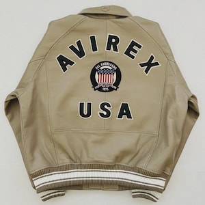 May include: Tan leather jacket with the word "AVIREX" across the back in black block letters. Below the word "AVIREX" is a circular patch with the words "ALL AMERICANS" and "USA" in black block letters. The jacket has a white and brown striped trim.