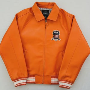 May include: Orange leather jacket with a collared neckline and a zipper closure. The jacket features a patch on the chest with the text "AVIREX" and an emblem. The cuffs and hem have white and red striped detailing.