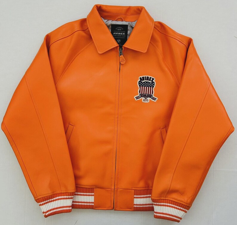 May include: Orange leather jacket with a collared neckline and a zipper closure. The jacket features a patch on the chest with the text "AVIREX" and an emblem. The cuffs and hem have white and red striped detailing.