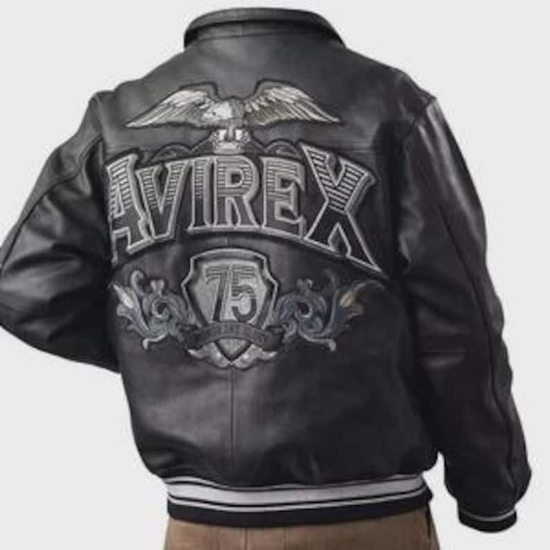 May include: Black leather bomber jacket with the word "AVIREX" embroidered across the back in silver and white. An eagle emblem is above the word, and a shield with the number "75" is below. The jacket has a ribbed collar and cuffs.