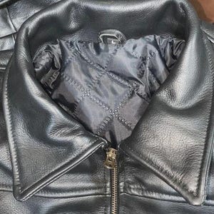May include: Close-up of a black leather jacket with a notched collar and a zipper closure. The jacket has a quilted lining and a patch on the lower right side. The jacket is a classic style.