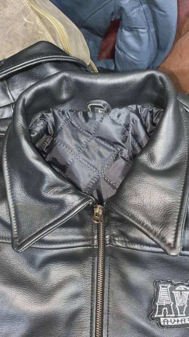 May include: Close-up of a black leather jacket with a notched collar and a zipper closure. The jacket has a quilted lining and a patch on the lower right side. The jacket is a classic style.