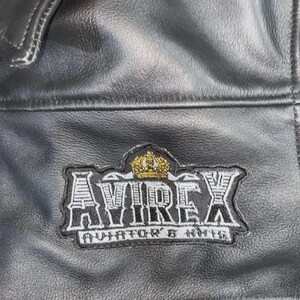 May include: Close-up of a black leather jacket with a stitched patch. The patch features the word "AVIREX" in a bold, stylized font, with a gold crown above and "AVIATOR'S KIT" below. The jacket has a textured surface.