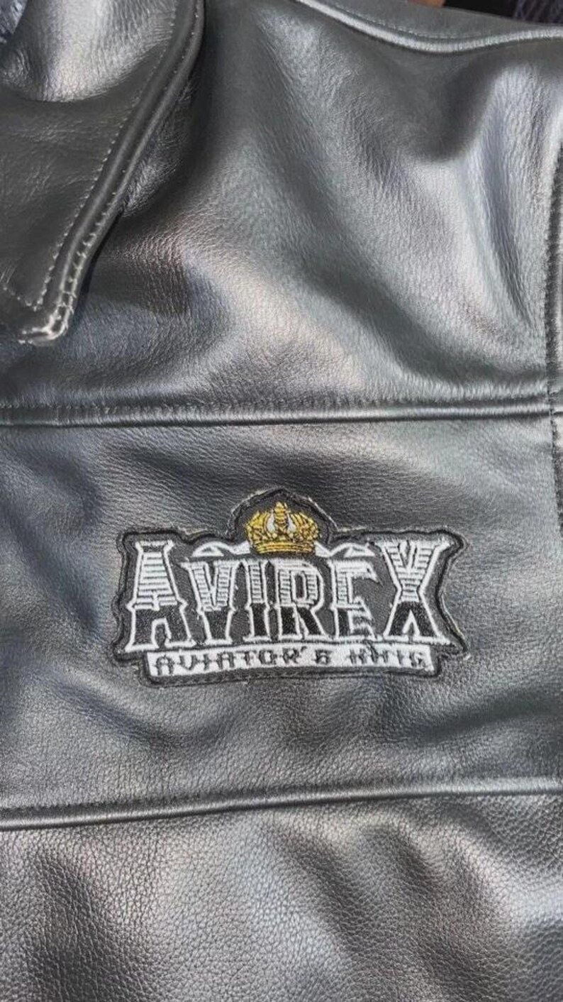 May include: Close-up of a black leather jacket with a stitched patch. The patch features the word "AVIREX" in a bold, stylized font, with a gold crown above and "AVIATOR'S KIT" below. The jacket has a textured surface.