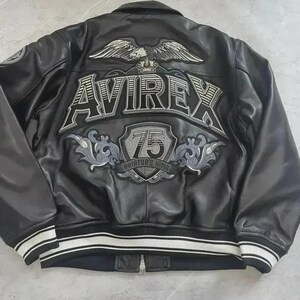 May include: Black leather jacket with white and black striped cuffs and hem. The back features the word "AVIREX" above a shield with "75 Aviator's King" and an eagle emblem. A classic bomber jacket style.