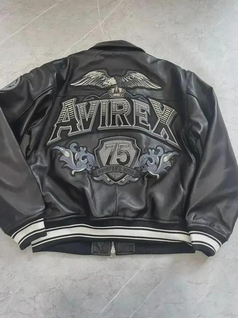 May include: Black leather jacket with white and black striped cuffs and hem. The back features the word "AVIREX" above a shield with "75 Aviator's King" and an eagle emblem. A classic bomber jacket style.