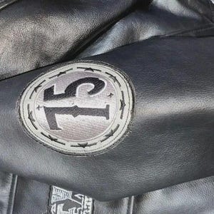May include: Black leather jacket with a circular patch on the sleeve. The patch features the number "75" in black against a gray background, surrounded by a gray border with star accents. The jacket has a classic design.