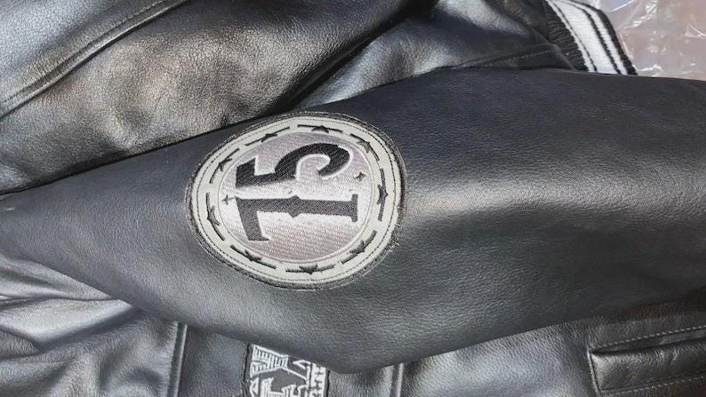 May include: Black leather jacket with a circular patch on the sleeve. The patch features the number "75" in black against a gray background, surrounded by a gray border with star accents. The jacket has a classic design.