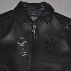 May include: Black leather jacket with a collared neckline and a zipper closure. The jacket features a patch on the chest and a tag with the brand name "AVIREX". The jacket is a classic style.