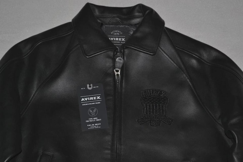 May include: Black leather jacket with a collared neckline and a zipper closure. The jacket features a patch on the chest and a tag with the brand name "AVIREX". The jacket is a classic style.