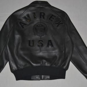 May include: Black leather jacket with the text "AVIREX USA" on the back. The jacket has a ribbed hem and cuffs, and a classic bomber jacket design. The jacket is a size large.