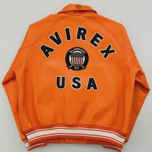 May include: Orange leather jacket with the word "AVIREX" across the back in black block letters. Below the word "AVIREX" is a circular patch with the words "ALL AMERICANS" and "USA" in black. The jacket has a white and black striped trim.