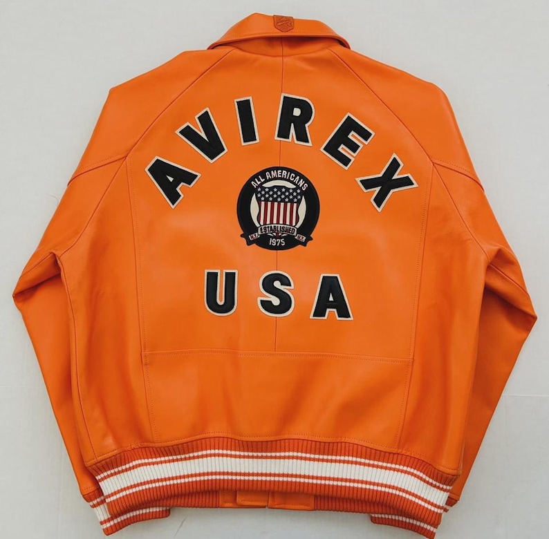 May include: Orange leather jacket with the word "AVIREX" across the back in black block letters. Below the word "AVIREX" is a circular patch with the words "ALL AMERICANS" and "USA" in black. The jacket has a white and black striped trim.