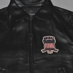 May include: Black leather jacket with a zipper closure and a collared neckline. The jacket features a patch on the chest with the text "AVIREX" above a design of red and white stripes and blue stars. The patch also includes the text "ESTABLISHED 1975".