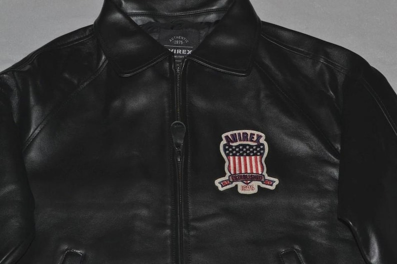 May include: Black leather jacket with a zipper closure and a collared neckline. The jacket features a patch on the chest with the text "AVIREX" above a design of red and white stripes and blue stars. The patch also includes the text "ESTABLISHED 1975".