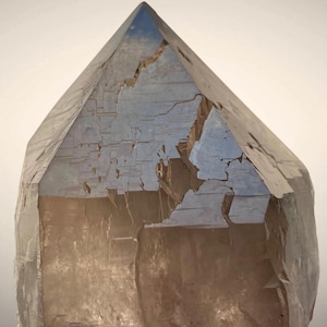 Può includere: Un grande cristallo sfaccettato con una parte superiore a punta. Il cristallo è di colore marrone traslucido con fratture interne visibili e una superficie leggermente riflettente. La forma del cristallo è una piramide alta e allungata.