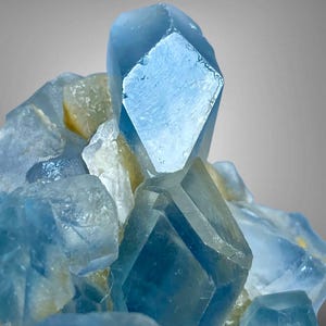May include: A cluster of translucent light blue crystals with sharp geometric facets and smooth surfaces. The mineral formation features a prominent central point surrounded by smaller, angular crystal structures with subtle pale yellow inclusions.