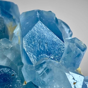 May include: A cluster of translucent, light blue fluorite crystals with sharp, geometric edges and smooth, glassy facets. The mineral formation displays varying shades of blue, ranging from pale sky blue to deeper azure, with small yellow inclusions.