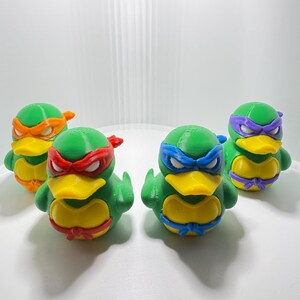 May include: Four green duck figurines, each with a different colored mask and bandanna, resembling the Teenage Mutant Ninja Turtles. The ducks have yellow beaks and bellies, with red, orange, blue, and purple accents.