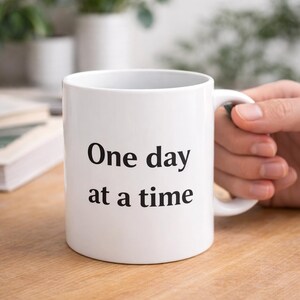 Pode incluir: Caneca de cerâmica branca com o texto "One day at a time" em preto. A caneca é segurada por uma mão e está sobre uma superfície de madeira. A caneca é cilíndrica com uma alça.