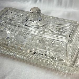 May include: A clear, rectangular glass butter dish with a lid and a tray. The lid has a small, round handle. The dish and lid feature a geometric pattern and are designed to hold butter. The tray has handles on each end.
