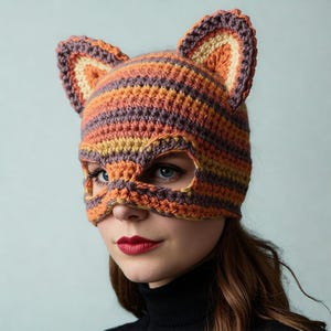 May include: A crocheted fox mask in shades of orange, purple, and yellow. The mask features ear details and eye openings. The handmade mask is worn by a person with red lipstick and a black turtleneck.