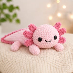 Könnte beinhalten: Ein handgefertigtes, rosa gehäkeltes Axolotl-Plüschtier mit dunkelrosa Kiemen und Akzenten. Das Axolotl hat schwarze Sicherheitsaugen und einen lächelnden Mund. Das Plüschtier liegt auf einer cremefarbenen Oberfläche.