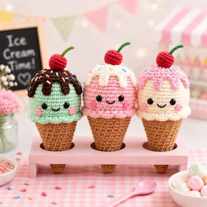 May include: Three crocheted ice cream cones with smiling faces, each topped with a red cherry. One is mint green with chocolate drizzle, another is pink with white sprinkles, and the last is cream with pink drizzle. They sit in a pink wooden holder.