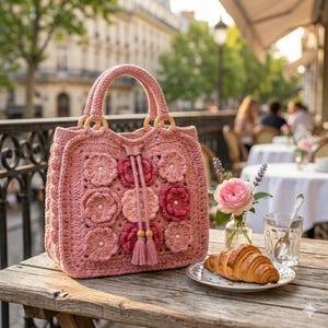 Peut inclure: Un sac à main rose en crochet avec un motif floral. Le sac est orné de poignées en bois et de pompons décoratifs. Il est posé sur une table en bois, avec une assiette de nourriture et un verre d'eau.