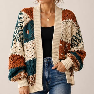 May include: A patchwork cardigan in cream, rust, and teal. The open-front sweater features a mix of knit textures and patterns, including cable knit and bobble stitch. The long-sleeved cardigan has ribbed cuffs and a cream-colored trim.