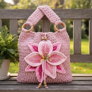 May include: A pink crochet handbag with wooden handles and a large, three-dimensional lily flower applique. The bag features a textured stitch pattern and is adorned with wooden beads. The bag is sitting on a wooden bench.