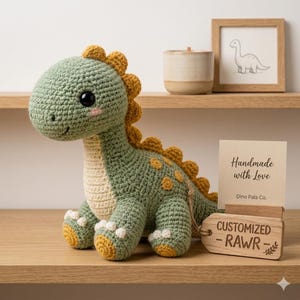 Puede incluir: Un peluche de dinosaurio tejido a ganchillo en verde salvia con detalles en amarillo mostaza en la espalda y el vientre. Tiene ojos de seguridad negros y mejillas rosadas. Una etiqueta de madera atada con cuerda dice "CUSTOMIZED - RAWR" junto a una tarjeta "Handmade with Love".
