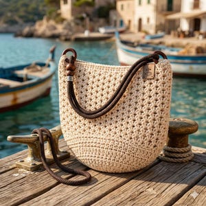 May include: A beige crochet tote bag with brown rope handles and wooden accents. The bag features a small rectangular tag with a fish design. The bag is sitting on a wooden dock near the water.