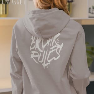 Könnte beinhalten: Ein hellgrauer Kapuzenpullover mit Kapuze und langen Ärmeln. Auf der Rückseite des Kapuzenpullovers befindet sich ein weißer Text im Graffiti-Stil mit der Aufschrift "BRUTE RULES". Der Text "PLATIN GREY" befindet sich oben links im Bild. Die Person trägt eine rosa Leggings.