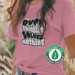 Könnte beinhalten: Ein rosafarbenes T-Shirt mit kurzen Ärmeln und dem schwarz-weißen Text "overthinking SOLVES nothing" in einer stilisierten Blasen-Schriftart. Ein kreisförmiges grün-weißes "ORGANIC 100 content standard" Logo ist unten rechts aufgedruckt.
