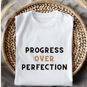 Op de afbeelding: Een wit t-shirt met de tekst "PROGRESS OVER PERFECTION" in zwart en bruin. Het shirt is opgevouwen op een geweven rieten dienblad, met gedroogde planten op de achtergrond.