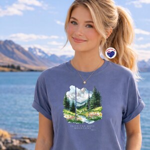 May include: A blue t-shirt featuring a mountain and river scene. The graphic shows trees, a stream, and a snow-covered mountain. The text "FAITH CAN MOVE MOUNTAINS" is below the image. The sleeves of the shirt are rolled up.