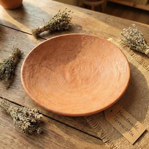 May include: A handmade terracotta bowl with a rustic, earthy tone. The bowl is surrounded by small bundles of dried herbs and a tag that reads "Handmade Terracotta". The bowl is approximately 25 cm in diameter.