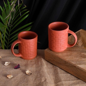 May include: Two terracotta-colored mugs with a brick-like texture. Each mug has a rounded handle and a cylindrical shape. They are displayed on a wooden tray and a burlap surface, with a dark background and some greenery.